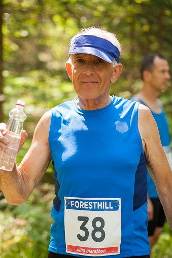 Senior male runner portrait in woods. People in the back. With numbers on dress. Drinking water. Preparing for start or just finnished.