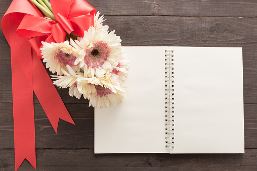 Pink gerbera flowers and notebook are in the wooden background with ribbon.