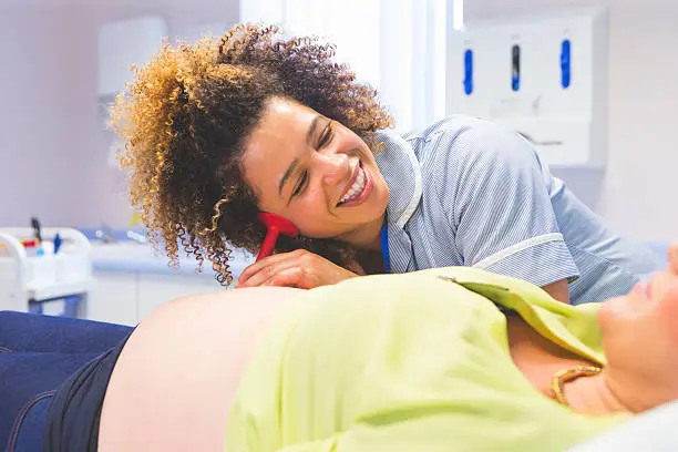 Doctor examining a pregnant woman. The woman is relaxed on the couch and the doctor has a pinard horn pressed to her stomach to listen to the baby's heartbeat.