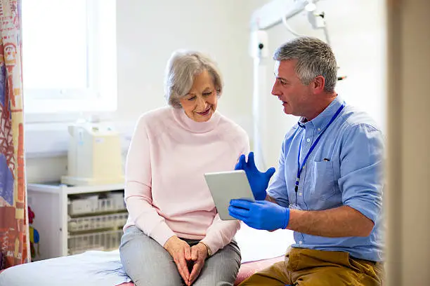 A male doctor sits beside an elderly female. They are in an examination room talking as he holds a digital tablet in one hand. The male doctor looks to be explaining something to his patient.