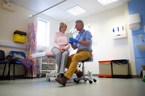 A male doctor sits beside an elderly female. They are in an examination room talking as he holds a digital tablet in one hand. The male doctor looks very happy as he smiles. View of room from a low angle