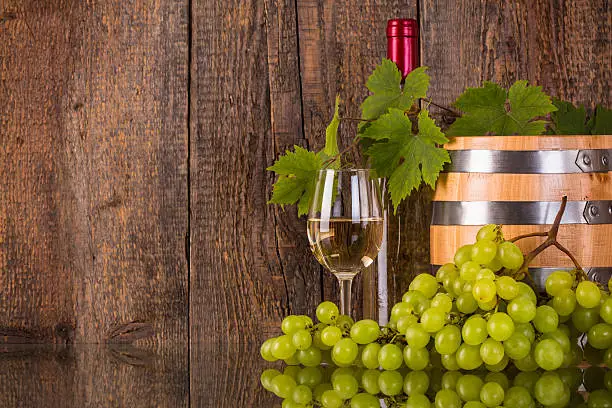 Glass of wine with a barrel white bottle hidden by grapeleaves and dark wooden background