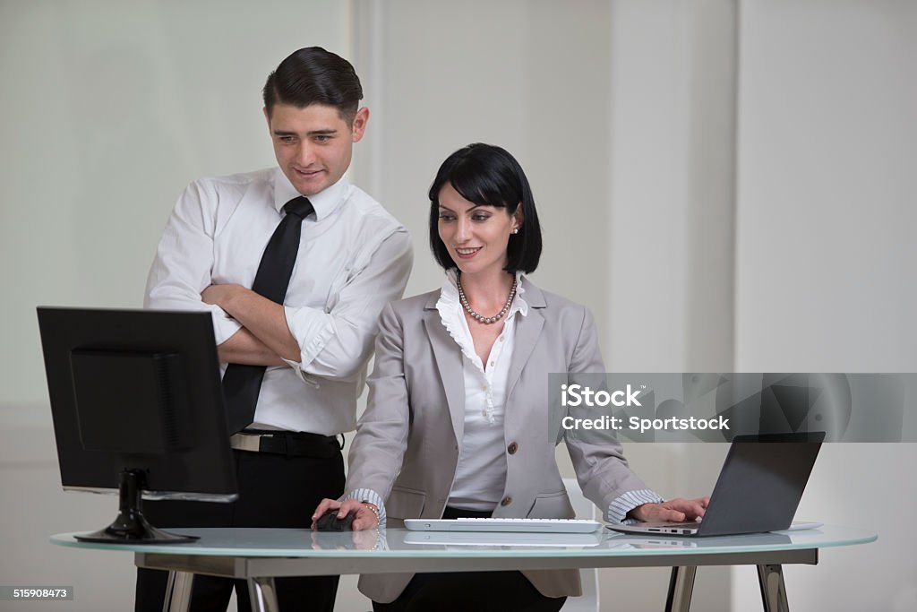 Two Executives Pleased With Their Work Horizontal photo of a businesswoman and a businessman working at a desk.  One person is standing and the other is sitting.  They are both looking at a computer.  The photo is set against a white, contemporary background.  20-29 Years Stock Photo