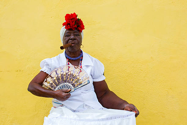 Havana, Cuba - March 28, 2015: Cuban lady wears traditional clothing with cigar and fan while sitting on a chair in the old town of Havana, Cuba.