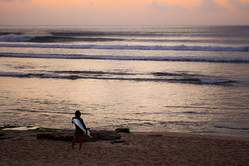 Balangan, Bali, Indonesia - August 9, 2014: The man is standing on the sand with his surfboard in front of the Indian Ocean. In background there are big perfect waves and the orange sky of the sunset.