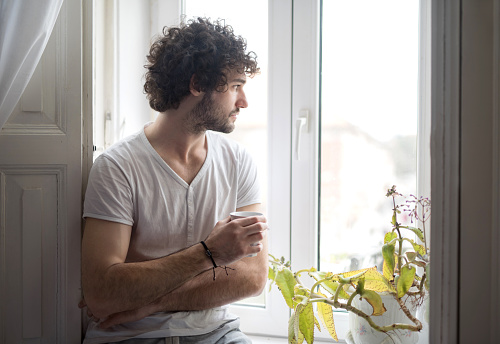 Young serious man at home thinking while holding cup of coffee and looking through the window