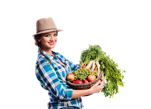 Beautiful young woman in blue checked shirt  and hat holding a basket full of vegetables and fruit. Studio shot on white background. Autumn harvest