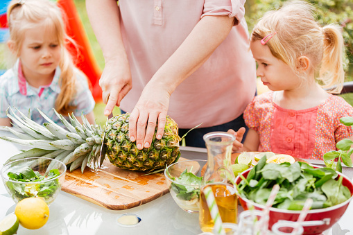 Two little girls preparing fresh smoothies outside. They are cutting fresh fruits and vegetables on wooden cutting board.