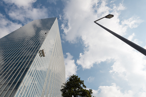 Modern office building against cloudy sky. The façades reflect blue sky and white clouds.  A gondola is hanging for window cleaning. Low angle view of the high rise building with a street lamp and a tree.