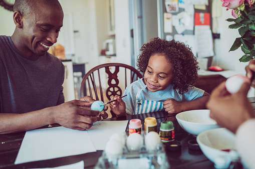 A photo of father and boy colouring Easter egg together. Happy man holding egg for son. They are sitting at table.