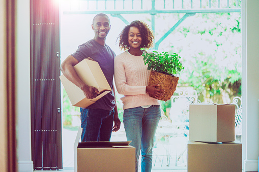 A photo of young couple with cardboard boxes and potted plant. Portrait of man and woman are moving house. They are smiling while standing at entrance of home.