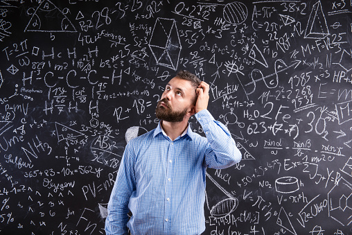 Hipster teacher scratching his head against big blackboard with mathematical symbols and formulas. Studio shot on black background.
