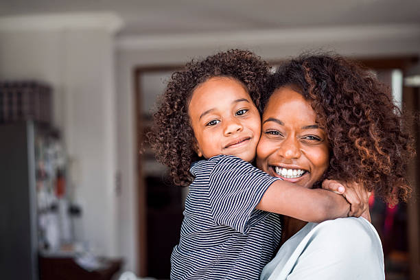 Happy mother and daughter embracing at home A photo of mother and daughter embracing. Portrait of happy woman and girl with curly and frizzy hair. They are in casuals at home. mom and baby eye contact stock pictures, royalty-free photos & images