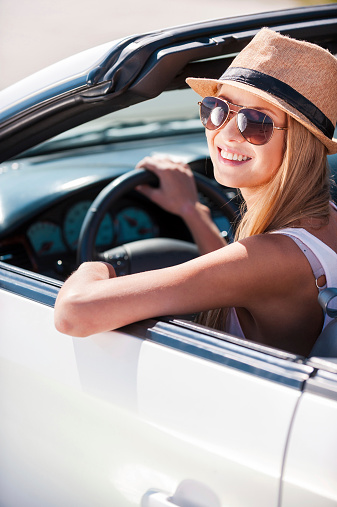Beautiful young funky woman in sunglasses sitting on front seat of her convertible and smiling