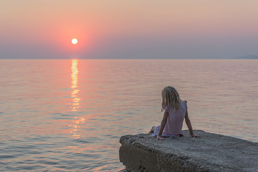 Young girl on her summer holiday gazing out to the setting sun on the horizon , it's warm colours reflected on the rippling sea water.