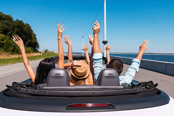 Rear view of young happy people enjoying road trip in their convertible and raising their arms up