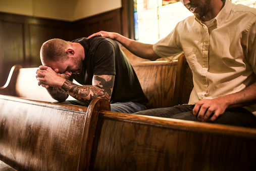 A man bows his head in prayer while a friend or pastor prays for him, his arm on his shoulder.  They sit on a wooden pew in a traditional church building, glowing stained glass windows visible behind them.  Horizontal image.