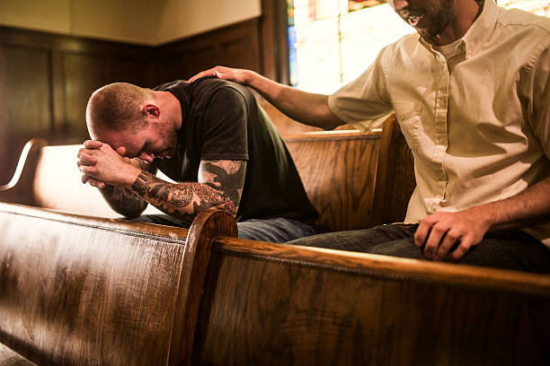Men Pray Together in Church A man bows his head in prayer while a friend or pastor prays for him, his arm on his shoulder.  They sit on a wooden pew in a traditional church building, glowing stained glass windows visible behind them.  Horizontal image. forgiveness stock pictures, royalty-free photos & images
