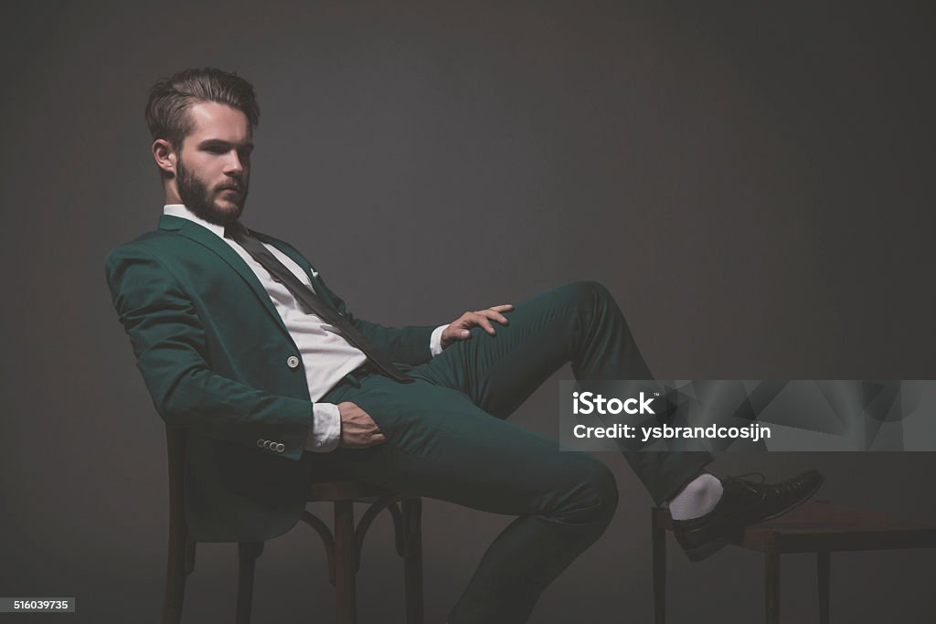 Fashion man wearing green suit with white shirt black a Business fashion man wearing green suit with white shirt black and tie. Sitting on wooden chair. Studio shot against grey. Adult Stock Photo