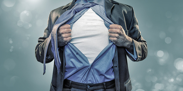 A conceptual, stylized close up image of a male torso dressed in business suit, shirt and tie, pulling open shirt to reveal a blank white t-shirt underneath in the manner of a superhero ready for action. This is a composite image, with a plain turquoise background and bokeh light effects.  
