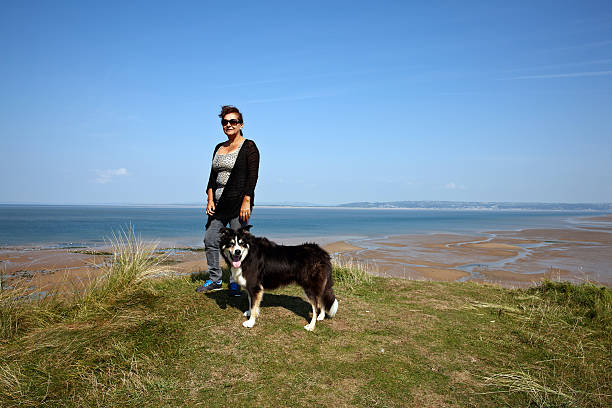 Mature hispanic woman standing on grassy headland and border collie is standing alongside