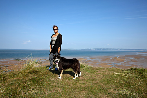 Mature hispanic woman standing on grassy headland and border collie is standing alongside