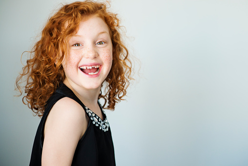 Portrait of a laughing little girl with flamboyant redhead and a missing tooth. She has curly hair and is looking at the camera. Light gray background. Focus on eyes. Horizontal indoors head and shoulders shot with copy space. This was shot in Quebec, Canada.