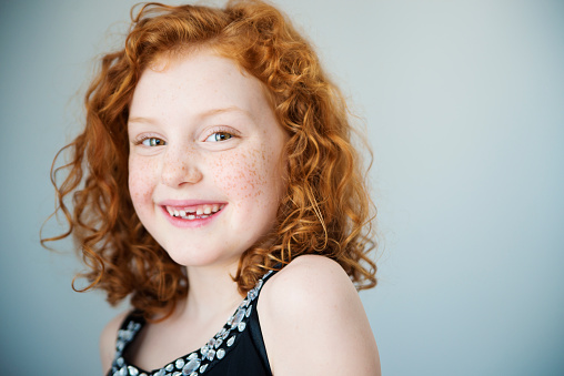 Portrait of a smiling little girl with flamboyant redhead and a missing tooth. She has curly hair and is looking at the camera. Light gray background. Focus on one eye. Horizontal indoors head and shoulders shot with copy space. This was shot in Quebec, Canada.