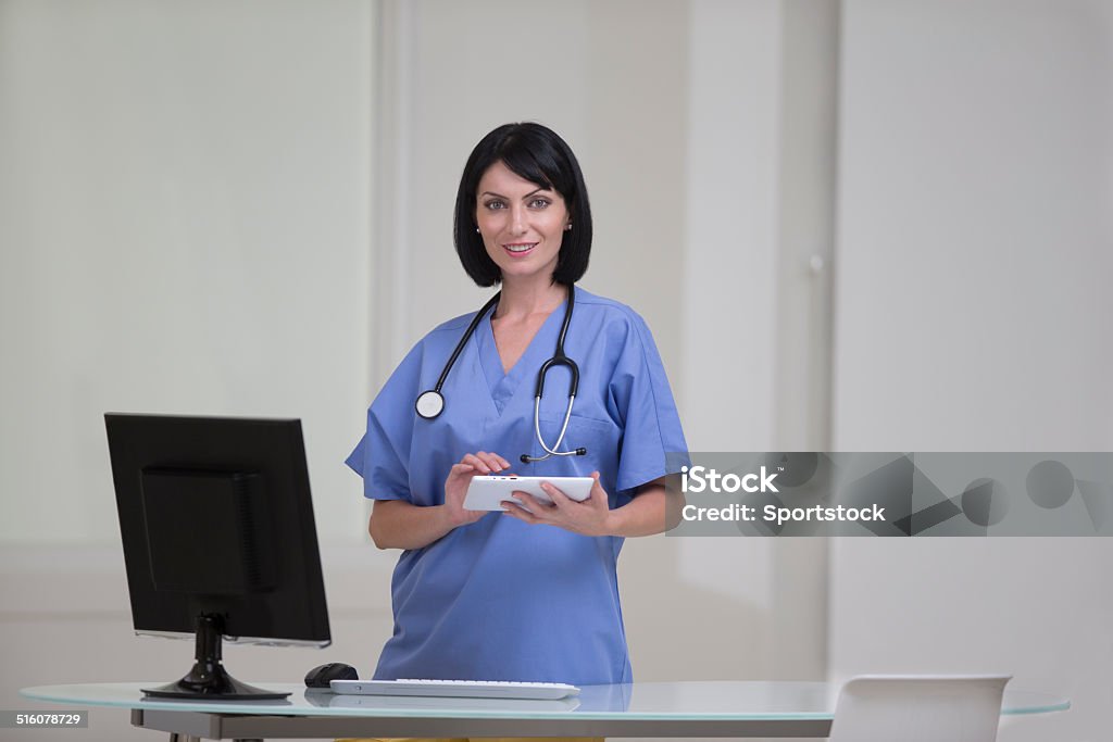 Portrait of a Doctor Standing at a Desk Horizontal photo of a beautiful young female medical professional standing at her desk and working on a digital tablet.  The photo is set against a white, contemporary background.  She is shown from the waist up.  There is only one person. 30-39 Years Stock Photo