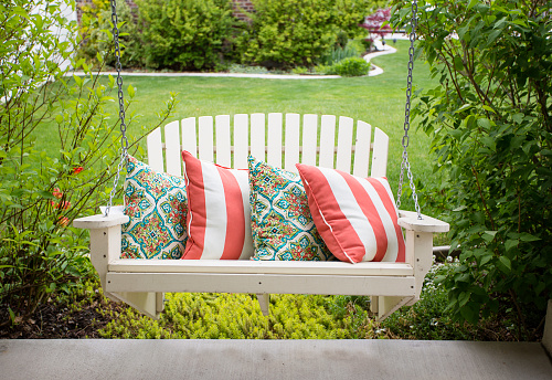 Beautiful wooden front porch swing with comfortable pillows. Outdoor porch seating with lots of green plants and shrubs around it.