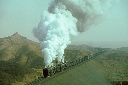 Two Chinese QJ class steam locomotives emit a huge cloud of exhaust smoke as they struggle uphill on a large embankment with a long line of loaded coal wagons and other freight in a remote and rugged desert region of Eastern China.  Horizontal, copy space, numbers and ID removed.
