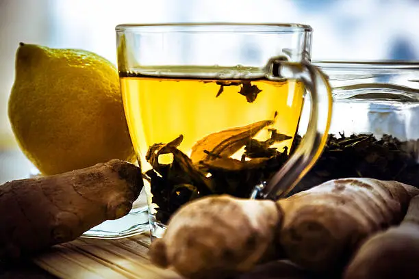 Lemon, ginger roots, glass jar with dry green tea and tea in a glass teacup on wooden table. Tea leaves are slowly sinking on to the bottom of te cup. Glass  teacup with   tea surrounded witg ginger roots and lemon, natural light.