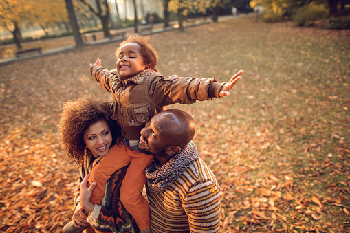 Happy African American family having fun in autumn park. Parents are holding their playful daughter on shoulders.