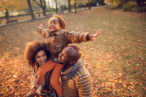 Playful African American little girl having fun with parents outdoors. Happy African American family having fun in autumn park. Parents are holding their playful daughter on shoulders. season stock pictures, royalty-free photos & images