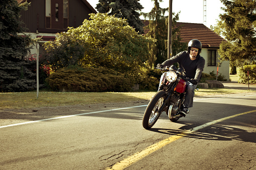 A handsome young adult male is riding a red vintage Cafe Racer styled Motorcycle around a bend wearing an open faced helmet, sunglasses and gloves. -- Post processing to look Vintage.  The man is wearing all black and the sun is low enough to cast alluring shadows.