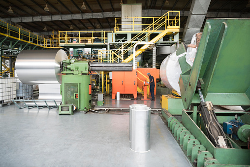 Factory worker using machine in aluminium can factory. Roll of aluminium sheet metal and machinery used in the processing of aluminium into drinks cans.