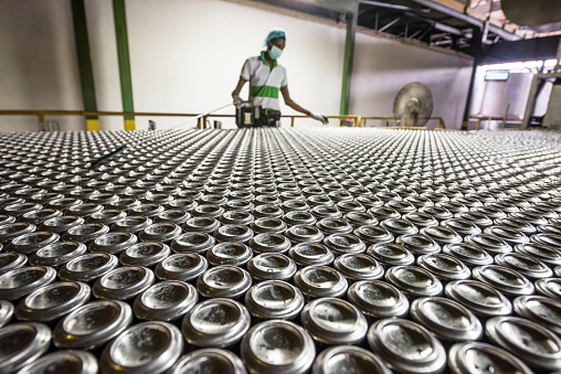 Aluminium drinks cans being produced in processing plant, male factory worker wearing mask working on production line.