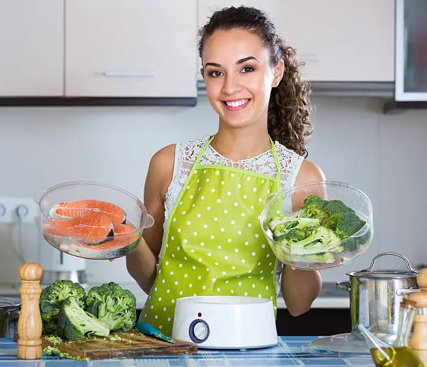 Smiling young brunette steaming salmon and vegetables in domestic kitchen