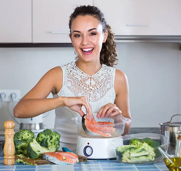 Cheerful young brunette woman steaming salmon and vegetables in domestic kitchen