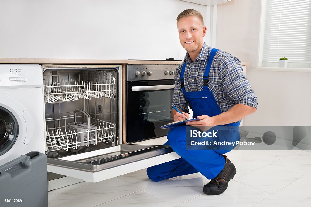 Handyman With Clipboard Looking At Dishwasher Young Handyman Looking At Dishwasher And Writing On Clipboard Adult Stock Photo