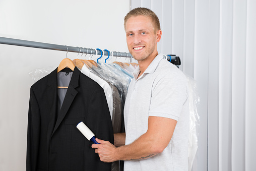 Young Happy Man Cleaning Dust With Lint Roller Of Coat In Clothes Store