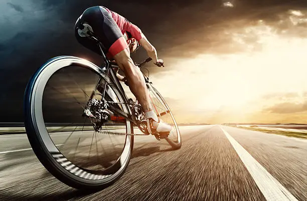 An athlete is riding a bicycle on road at the evening. The man is wearing black bike shorts and shin guards along with a red sleeveless top and a red and white helmet and sunglasses. The image is blurred in motion.