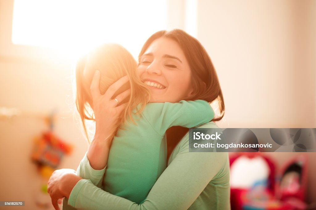 Mother and daughter hugging Mother is hugging her daughter at home. Bright light is coming through the window in the background. Mother Stock Photo