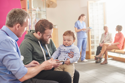 a mature male doctor sits next to a male patient and his baby son in his waiting room and casually chats to the father  . In the background a nurse chats to a mother and grandmother with her clipboard.