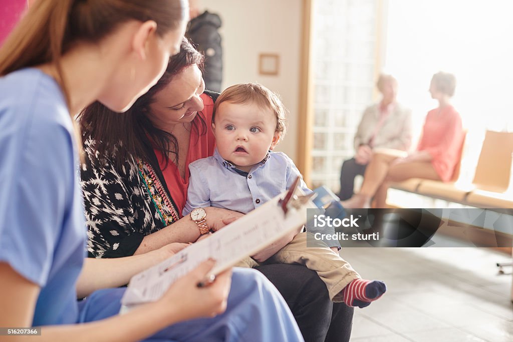 baby clinic visit for toddler and mum a female nurse sits next to a woman and her  baby son in the waiting room of a clinic and casually chats to them  . In the background two patients sit and chat to a senior doctor . Busy Stock Photo