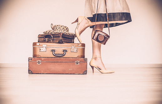 Woman standing with her vintage suitcases inside a home concept symbolizing she is going away on a trip or going on a vacation. Woman is  wearing a beige dress and shoes. She is holding a vintage camera. Only showing the woman's legs, hands and shoes. Horizontal sepia image. Copy space.