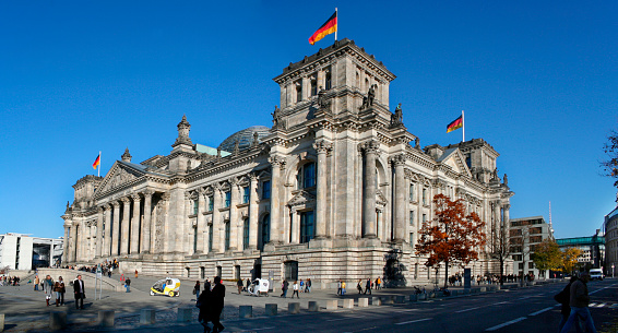 Berlin, Germany - March 17, 2016. Panoramic diagonal front view of the Bundestag, on a sunny autumn afternoon, with several German flags waiving on top of the building against a deep blue sky.  Visitor are walking around or waiting on the stairs of the building. 