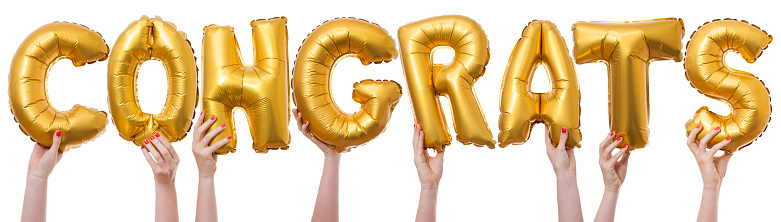 The word congrats has been created by female hands holding individual letter balloons. Each balloon is being held against a plain white background by a caucasian female hand. The Balloons are inflated and made from a shiny reflective gold material.