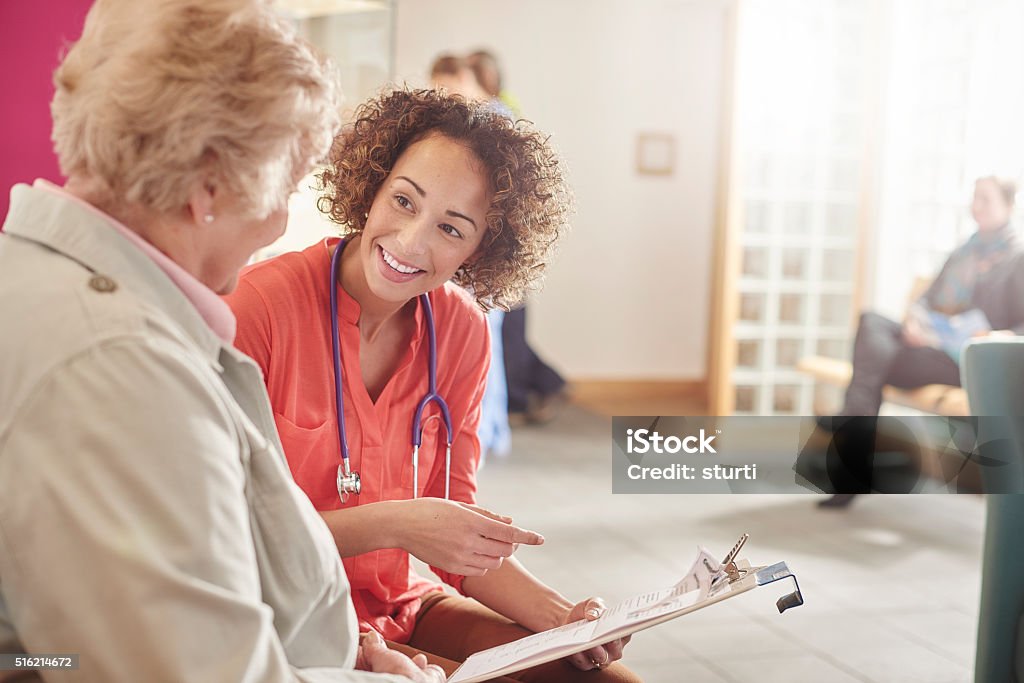 happy health senior with GP a  female doctor stands next to a senior patient in her waiting room and casually chats to her about her details . In the background a nurse chats to woman with her digital tablet , and a male patient stands at the reception desk and booking an appointment . Patient Stock Photo