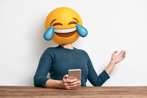 Emoji Head Woman sitting at desk. Woman wearing tears of joy emoji masks while looking at her phone. This emoji is laughing so much that it is crying tears of joy